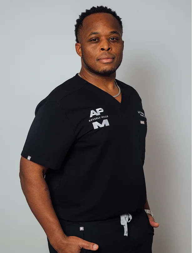 A studio portrait of Dr. Marshall against a light grey background. He is wearing black medical scrubs with "AP Beverly Hills" and "A.P. Marshall, MD Plastic Surgery" embroidered on the chest.