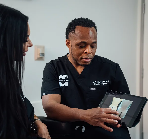 Dr. Marshall, wearing black scrubs with "A.P. Marshall, MD Plastic Surgery" branding, conducts a consultation with a female patient. He is using a tablet to point out specific details on "before and after" body contouring results.