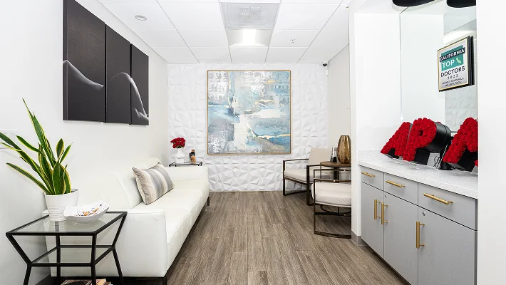 The interior of a modern, luxury waiting room. It features a white leather sofa, a large abstract blue and gold painting on a textured 3D geometric wall, and minimalist black side tables with magazines.