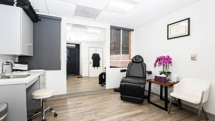 A view of a clean, modern clinical treatment room. The room includes a black adjustable medical chair, a sink area with grey cabinetry, a white guest chair, and a side table topped with a vibrant purple orchid. A framed diploma hangs on the white wall.