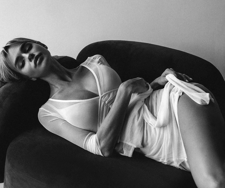 A black-and-white artistic photograph of a woman reclining on a dark velvet sofa, wearing a sheer white garment.