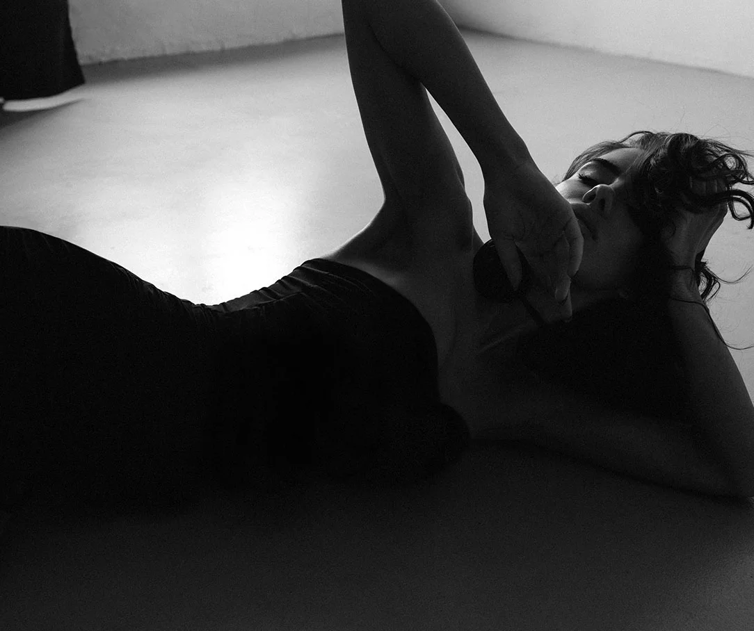 A black and white photo of a woman in a dark strapless top lying on a smooth floor with one arm raised over her face.