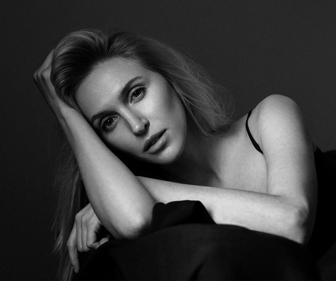 A black and white portrait of a woman resting her head on her arms and looking directly into the camera.