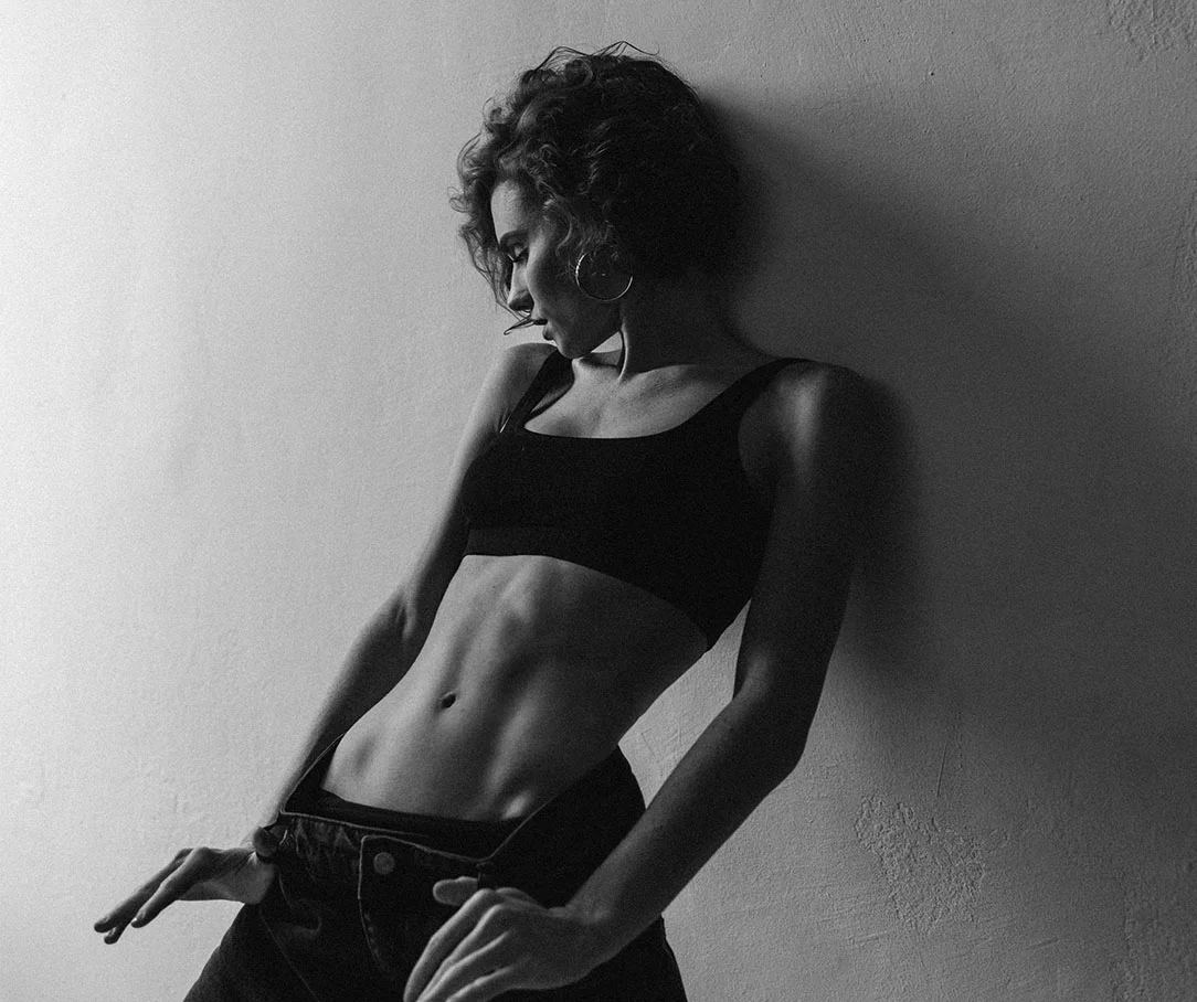 A black and white side-view of a woman in a black crop top and jeans leaning against a wall, showcasing a toned midsection.
