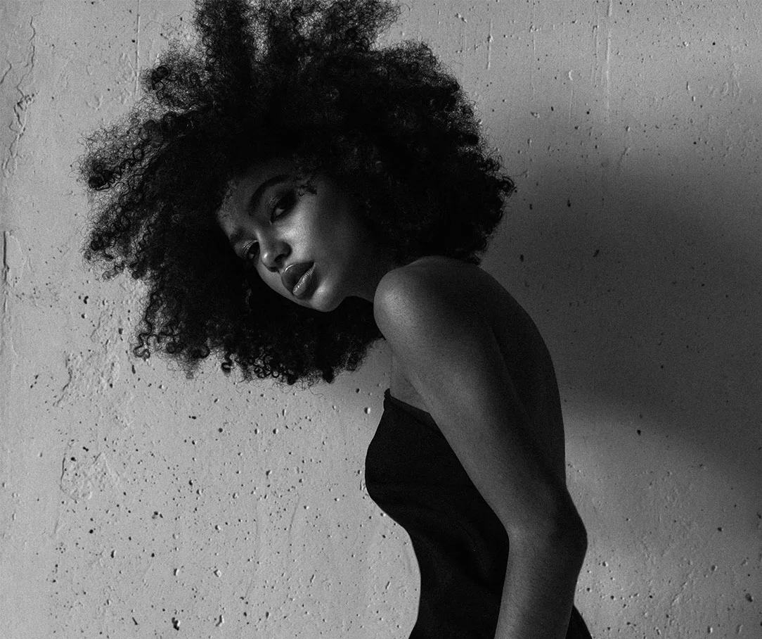 A dramatic black and white photograph of a woman with voluminous natural hair, posing in profile against a textured concrete wall.