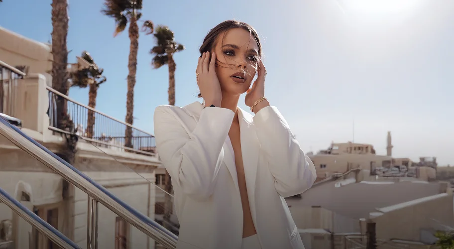 A high-fashion portrait of a woman in a white blazer against a bright, sun-lit urban rooftop background with palm trees. She has her hands near her face, highlighting her defined facial features.