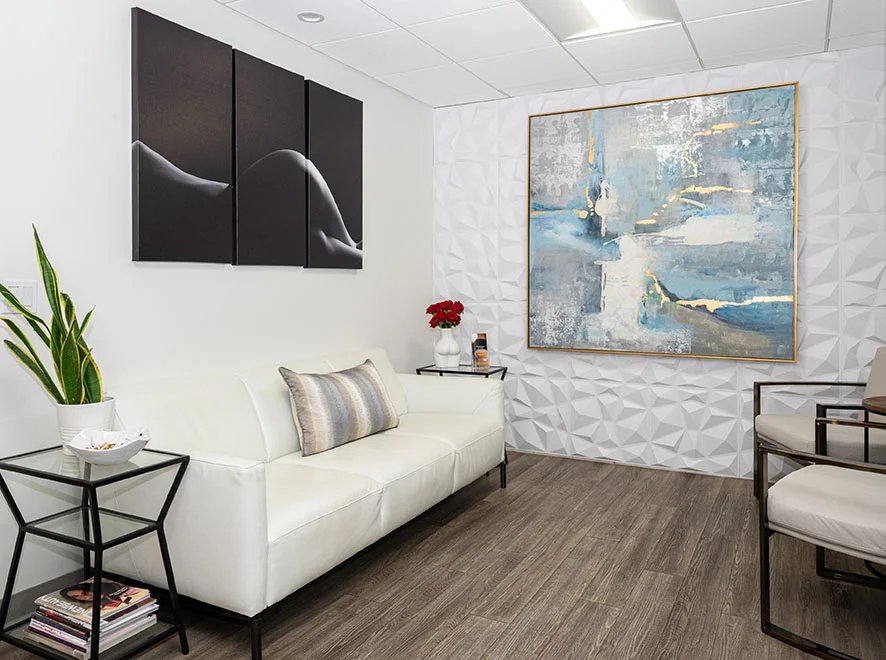 The interior of a modern, luxury waiting room. It features a white leather sofa, a large abstract blue and gold painting on a textured 3D geometric wall, and minimalist black side tables with magazines.