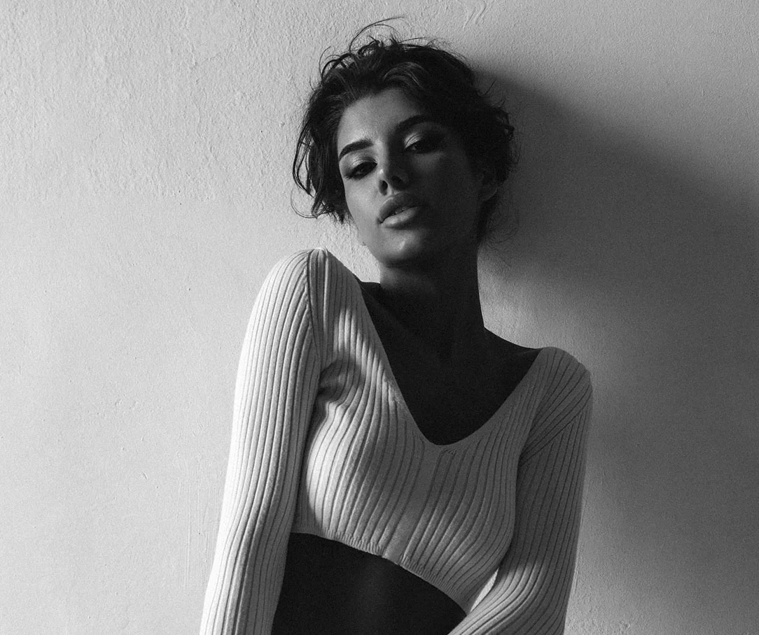 Black and white photo of a woman in a white ribbed cropped sweater leaning against a white wall.
