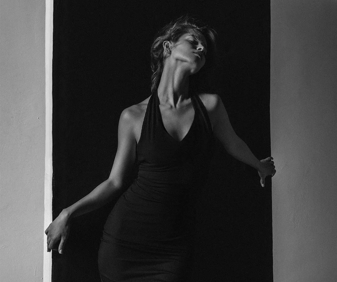 Black and white silhouette of a woman in a black halter dress posing against a high-contrast wall.