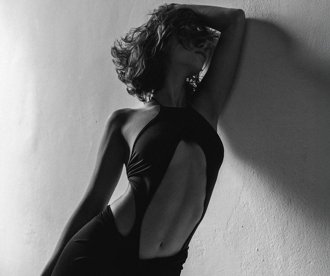 A black and white artistic photo of a woman in a cut-out black bodysuit leaning against a wall, showcasing her midsection.
