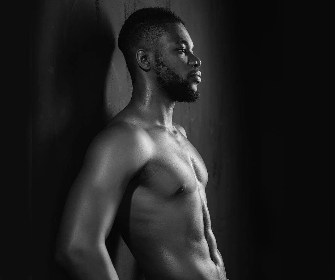A black and white profile shot of a shirtless man with a beard leaning against a dark wall, highlighting his athletic build.