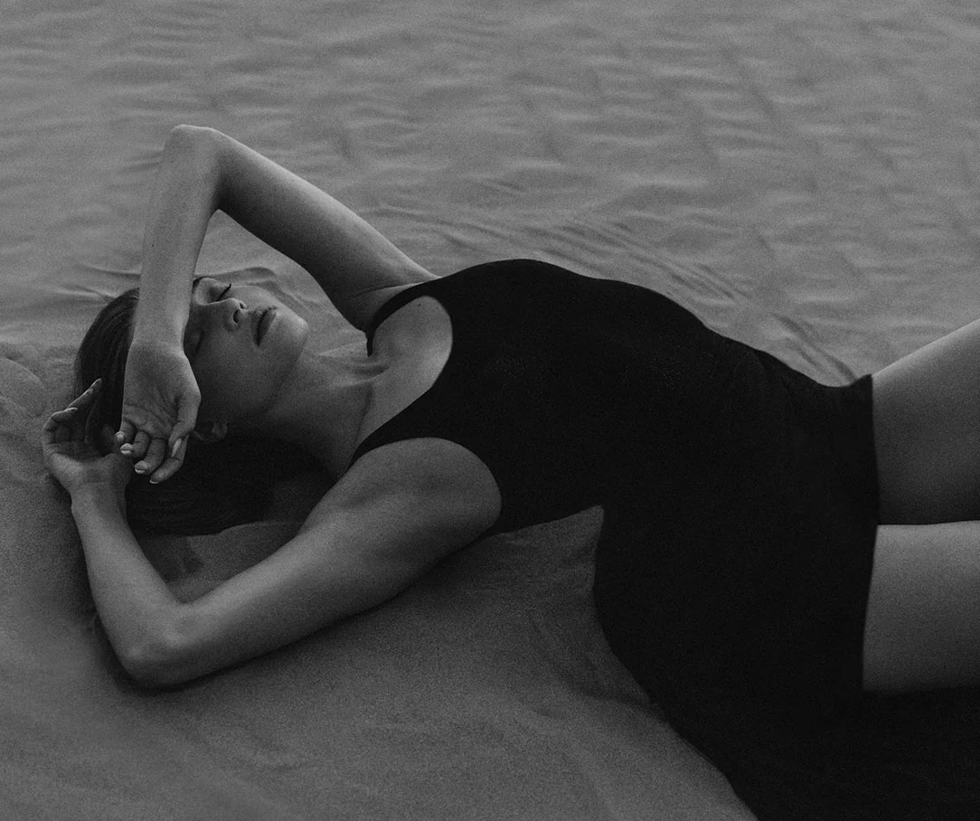A black and white photo of a woman reclining on sand with her arm over her forehead, wearing a black swimsuit.