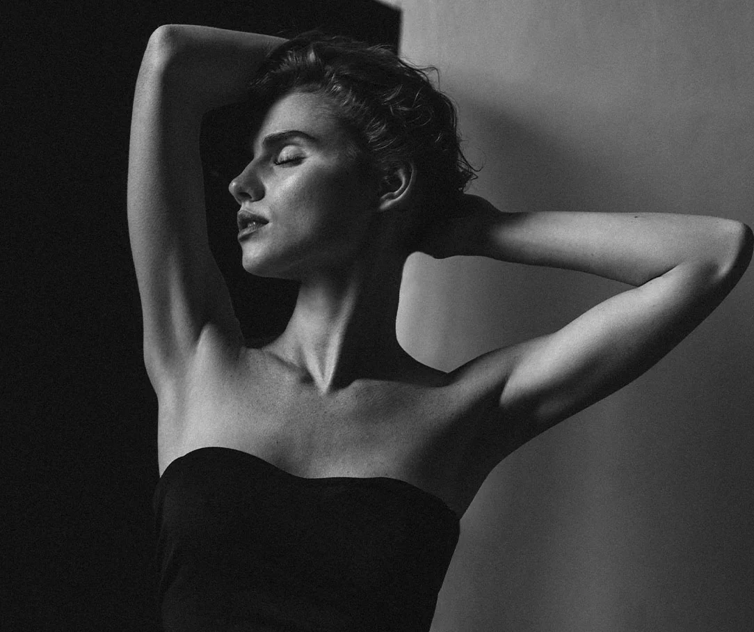 Artistic black and white photo of a woman in a black strapless top with her arms raised behind her head.